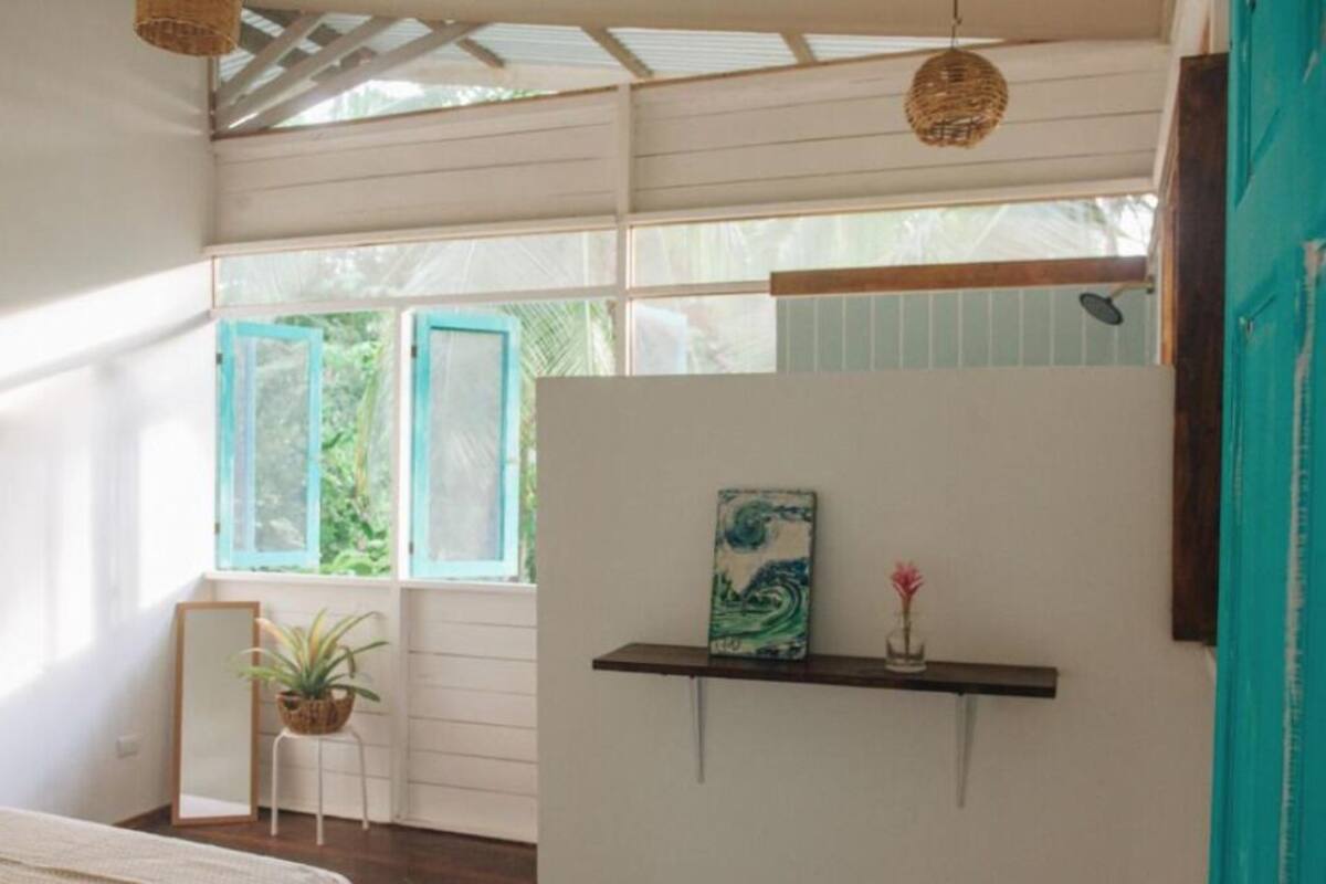 Una habitación con una cama, una mesita con una planta, un estante en la pared con un cuadro y una ventana con contraventanas azules.