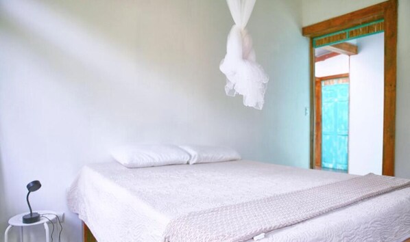 A bedroom with a bed, white pillows, a white blanket, a bedside lamp, and a wooden door with a green frame. - CASA BOHÖ (Cahuita)