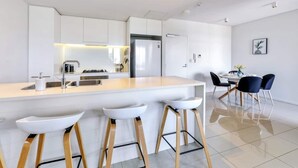 A modern kitchen with a central island, bar stools, a dining area with chairs, and a refrigerator. - Waterfront Sunset Suite: 1 Bed Apt, Sunset Views (Darwin City)