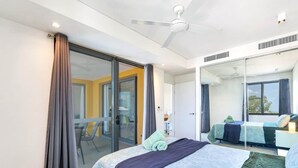 A modern hotel room with a large bed, a sliding glass door leading to a balcony, a ceiling fan, and a large mirror reflecting the room. - Waterfront Sunset Suite: 1 Bed Apt, Sunset Views (Darwin City)