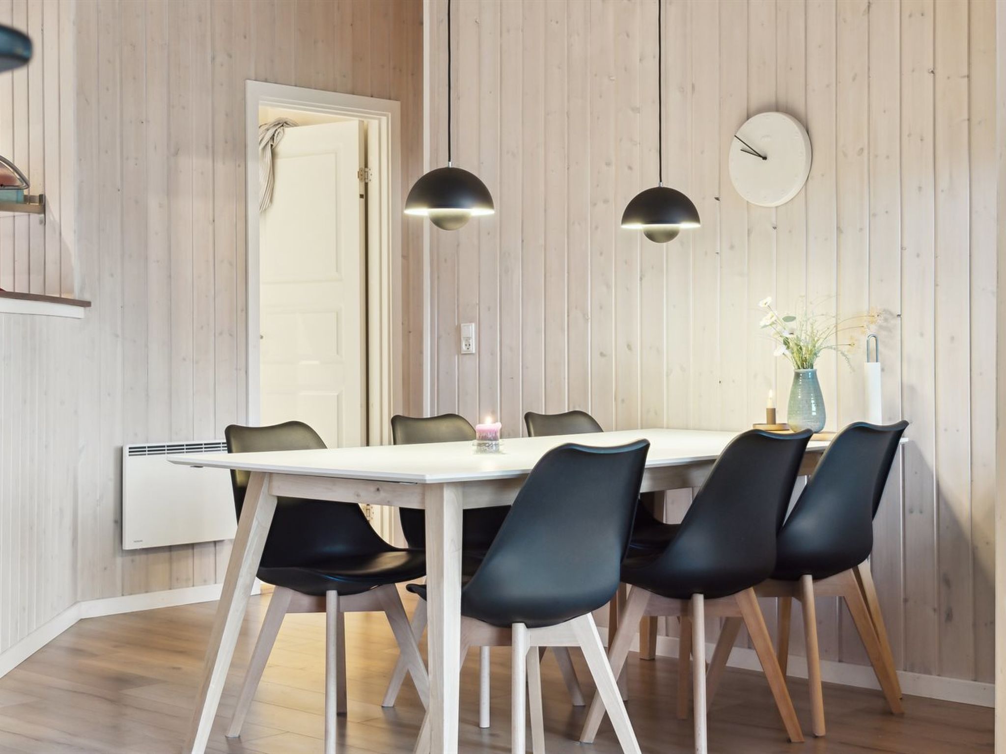 A dining area with a white table, black chairs, pendant lights, and a clock on the wall.