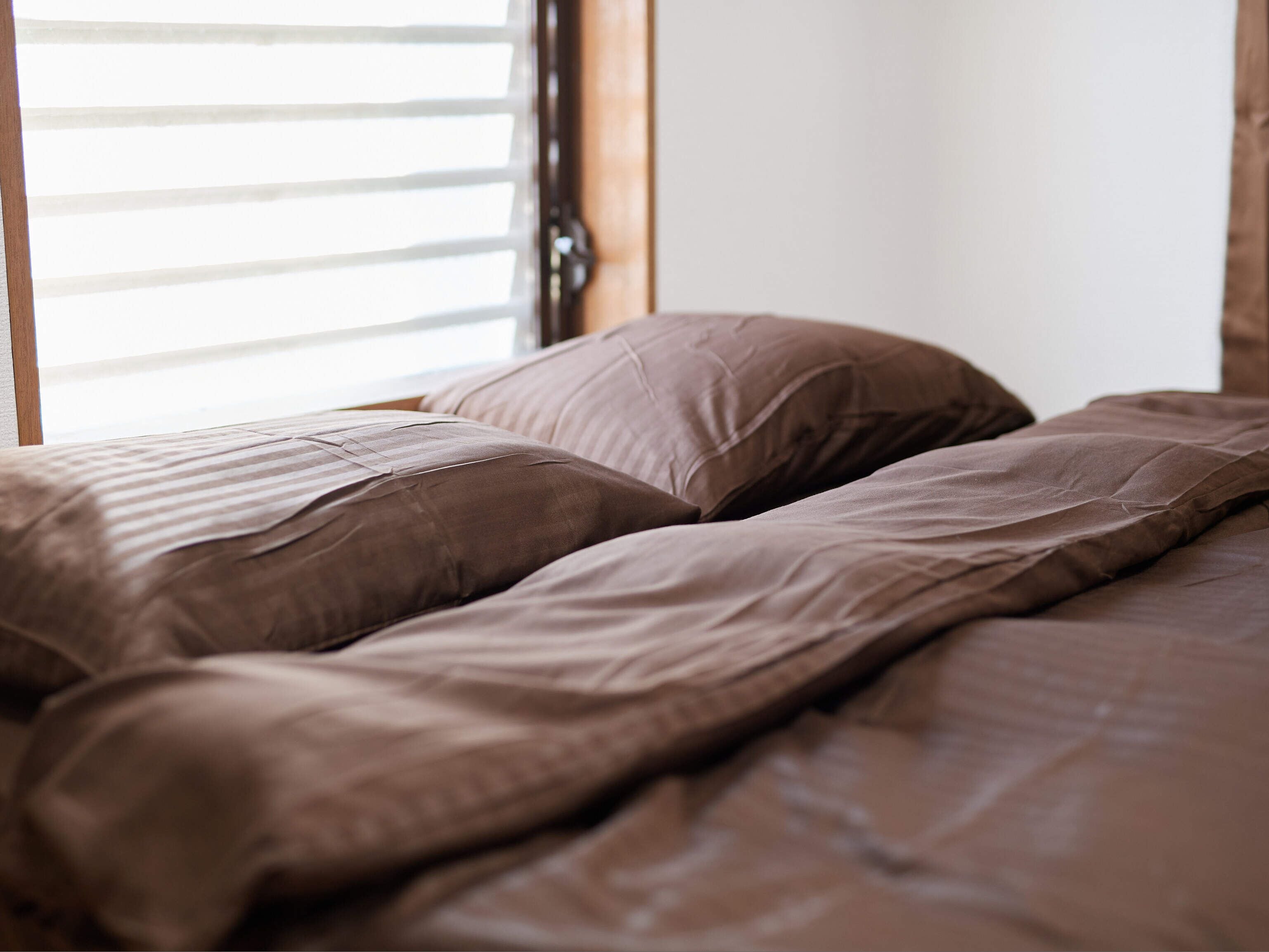 Una cama con una manta marrón y almohadas, una ventana con persianas, y un cabecero de madera.