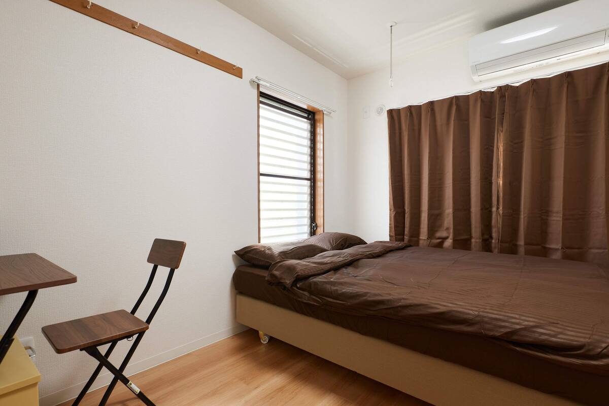 A small, neatly arranged bedroom with a bed, a chair, a table, and a window with blinds.