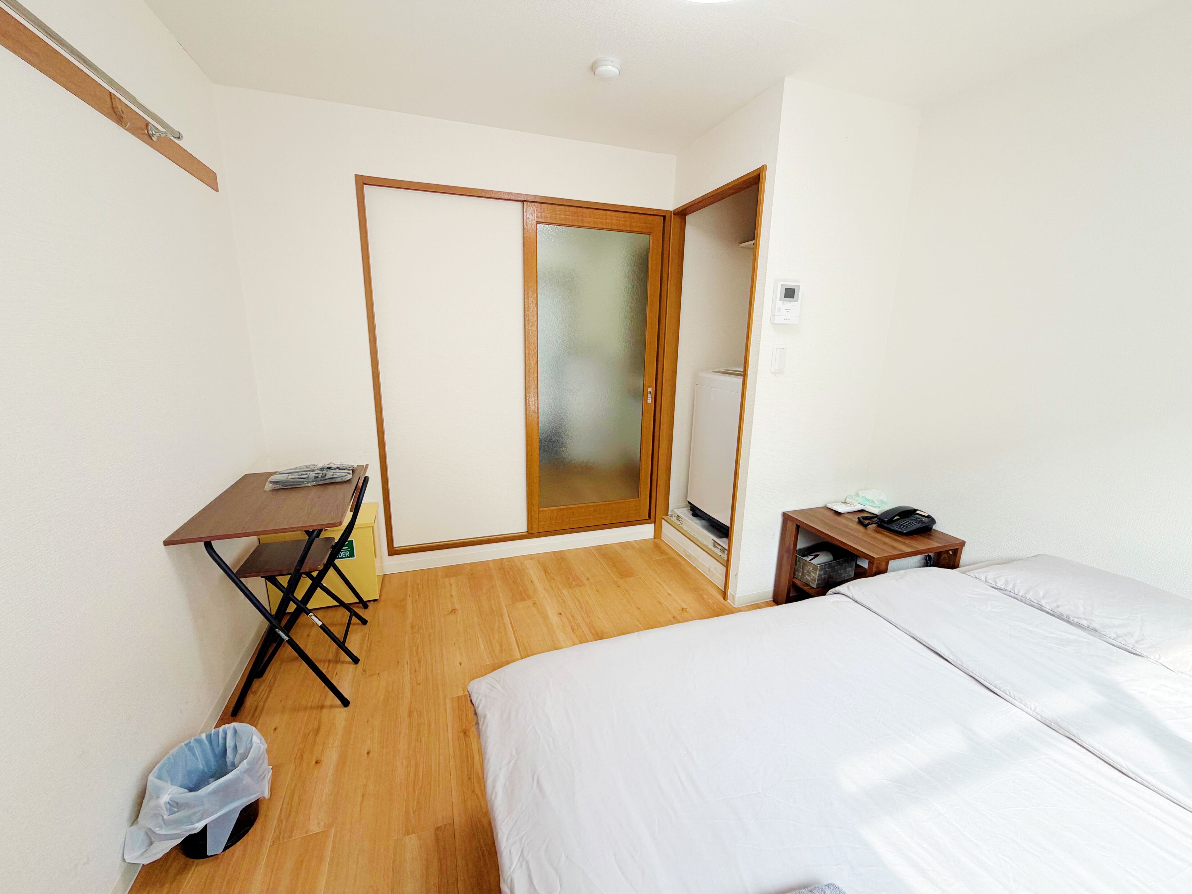 Een slaapkamer met een bed, een bureau, een stoel, een klein tafeltje met een telefoon, een prullenbak en een kledingkast met schuifdeur.