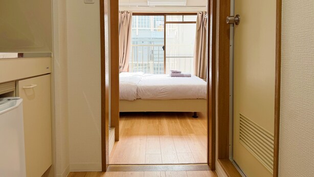 Ein schmaler Flur, der zu einem Zimmer mit einem Bett, einem mit Vorhängen versehenen Fenster, einer Deckenleuchte und einer Wand-Klimaanlage führt.