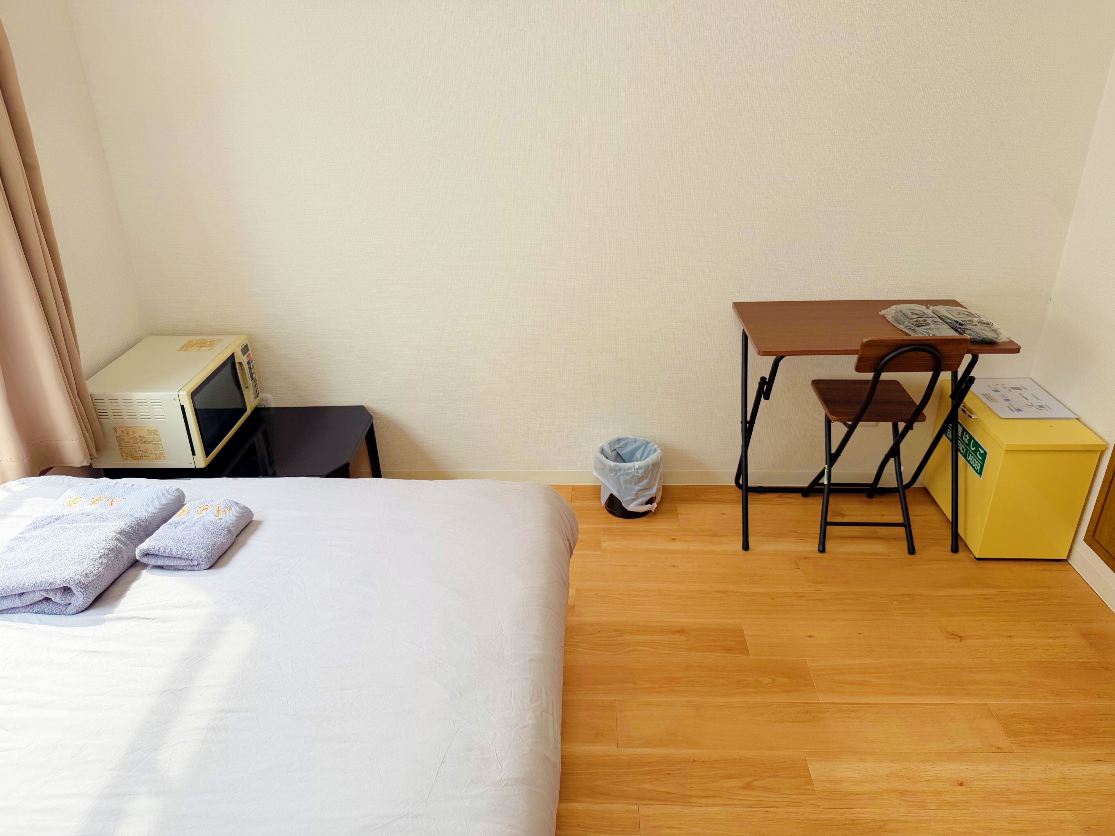 A hotel room with a bed, a desk, a chair, a microwave, a trash bin, and a yellow box.