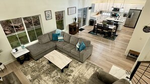 Un vaste espace de vie comprenant un grand canapé d’angle, une table basse et une table à manger avec des chaises. Au fond, on trouve une cuisine équipée d’appareils électroménagers modernes et un coin repas.