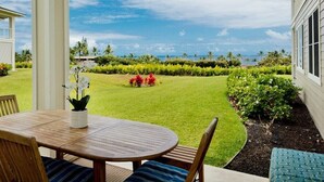 Um pátio com mesa e cadeiras de madeira, um pequeno jardim com diversas plantas e flores, e vista para o oceano e palmeiras.