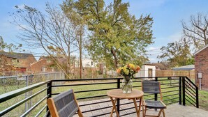 Ein Balkon mit Tisch und zwei Stühlen, einer Blumenvase und Blick auf ein Wohngebiet mit Häusern und Bäumen.