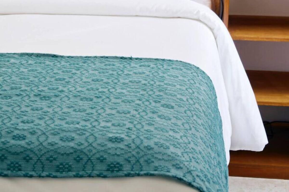 A neatly made bed with white linens and a teal patterned throw, a wooden headboard, and a small bedside table with a woven lamp.