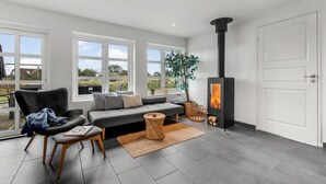Uma sala de estar moderna com uma lareira a lenha preta, um sofá cinza, uma poltrona preta e janelas amplas com vista para o exterior.