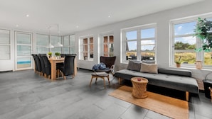 Uma sala de estar ampla com um sofá cinza, uma mesa de jantar com cadeiras e grandes janelas com vista para o exterior.