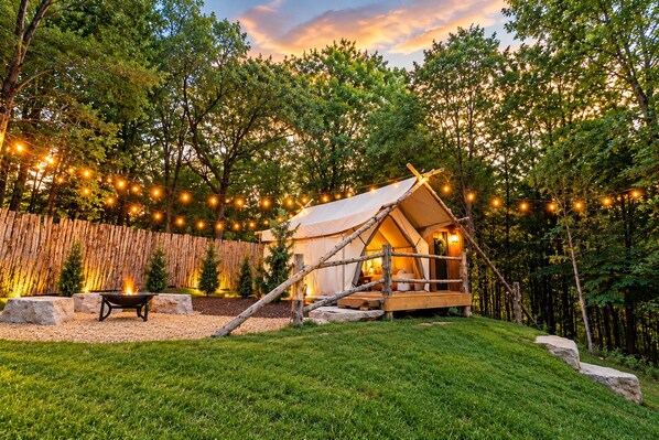 A rustic tent with a fire pit and string lights in a wooded area.