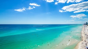 Uma praia com águas turquesa cristalinas, um cais e o horizonte de uma cidade ao fundo.