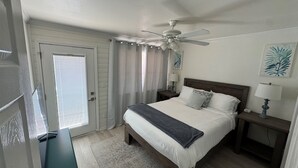 Un dormitorio con cama, ventilador de techo, mesita de noche con lámpara y puerta con cortinas.