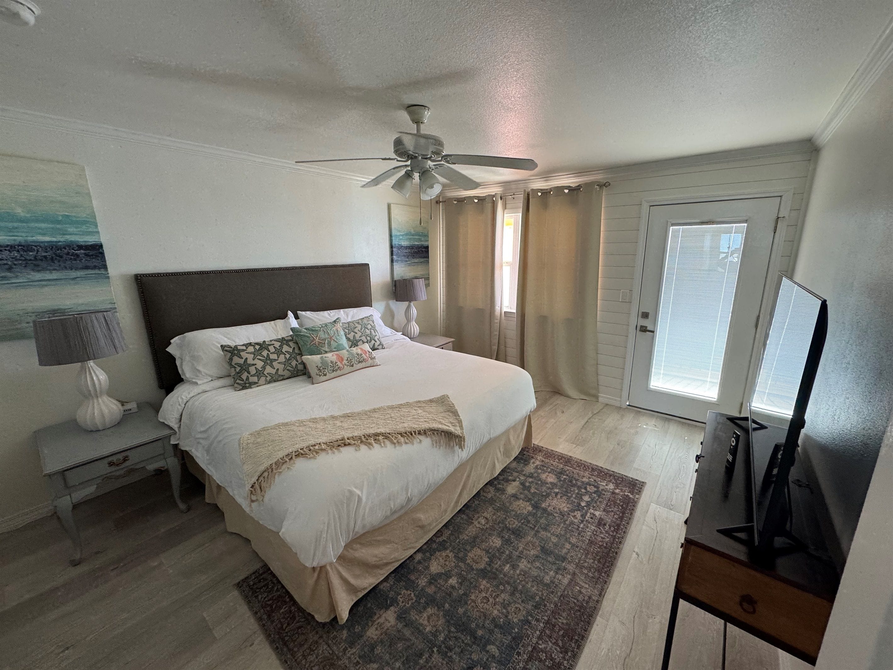 Un dormitorio con cama, mesitas de noche, ventilador de techo, televisor y ventana con cortinas.