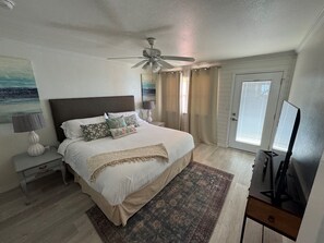 Un dormitorio con cama, mesitas de noche, ventilador de techo, televisor y ventana con cortinas.