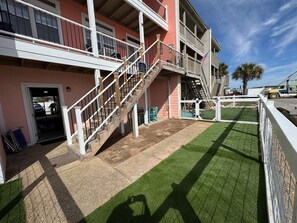 Un edificio de varios pisos con una escalera que lleva a un balcón, un patio cercado con césped artificial y un cielo despejado.