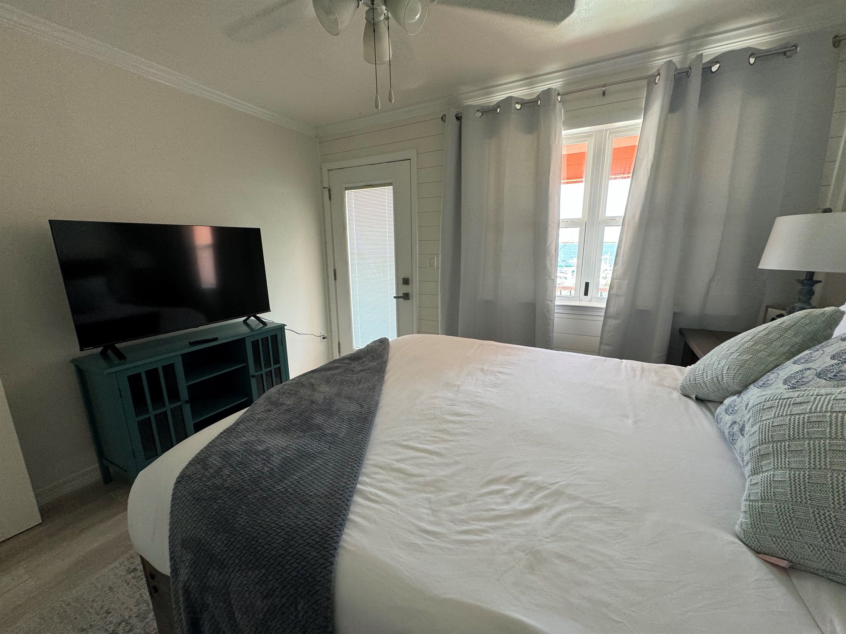 A bedroom with a bed, a television on a stand, a window with curtains, and a ceiling fan.