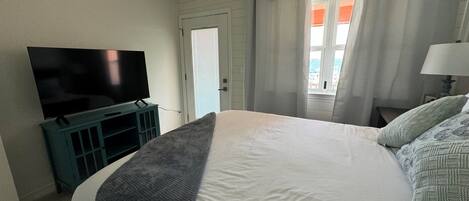 A bedroom with a bed, a television on a stand, a window with curtains, and a ceiling fan.