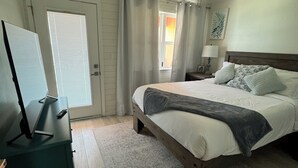 Un dormitorio con cama, ventilador de techo, mesita de noche con lámpara, escritorio con televisor y ventana con cortinas.