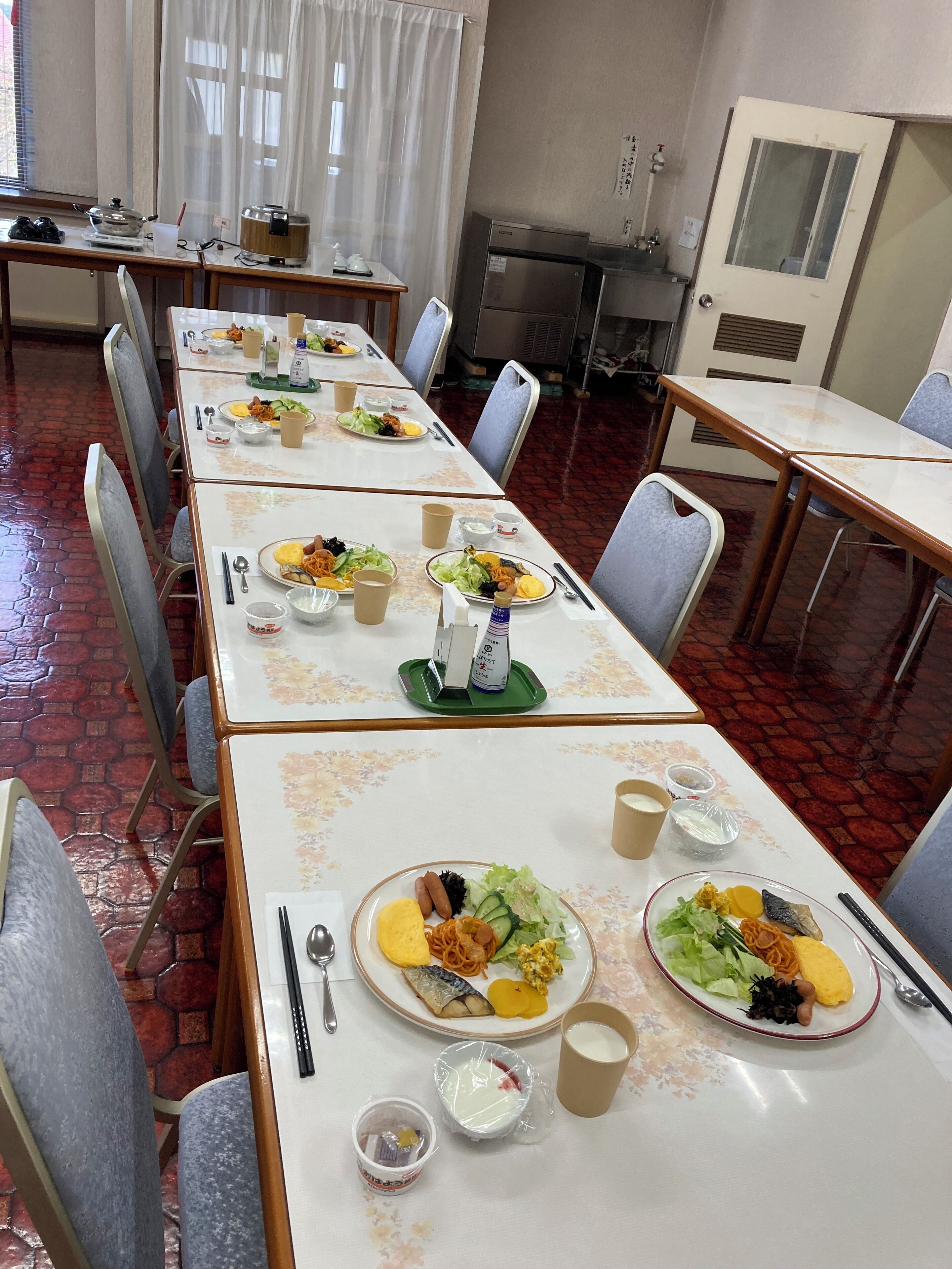 A dining area with multiple tables set for meals, chairs, and a kitchen area in the background.