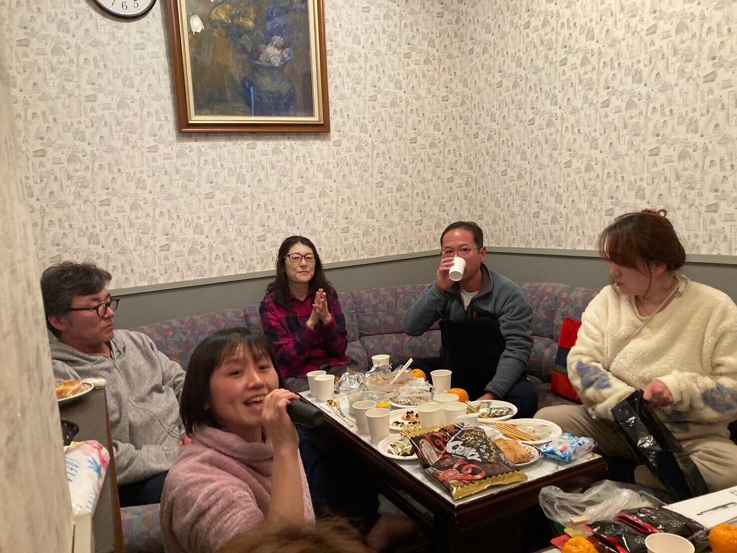 Un grupo de personas sentadas alrededor de una mesa con comida y bebidas, en una habitación con papel tapiz estampado y un reloj en la pared.