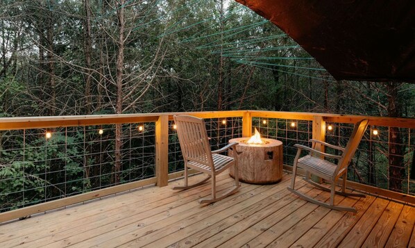 A wooden deck with two rocking chairs and a central fire pit, surrounded by a wire mesh fence and string lights. - Treehouse Branson, MO Cozy Glamping Site (Branson)