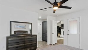 Une chambre à coucher comprenant un lit, une commode, un ventilateur de plafond et une porte donnant sur une autre pièce.