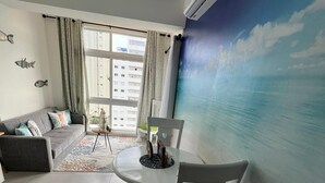 Uma sala de estar moderna com um sofá cinzento, uma mesa de jantar redonda com cadeiras brancas e uma grande janela com cortinas.