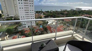 Varanda com vista para o oceano, uma cadeira, uma pequena mesa e um edifício branco ao fundo.