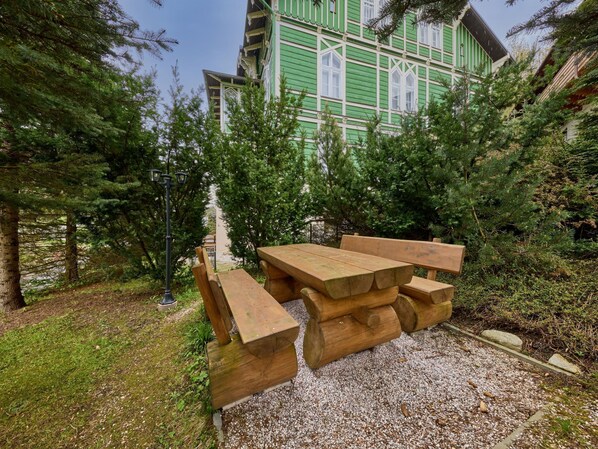 Una mesa de picnic de madera con bancos en un jardín con árboles y un edificio al fondo.