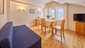 A cozy room with a wooden floor, a sofa, a dining table with chairs, a TV, and a window with a view of trees.
