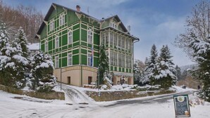 A large, multi-story building with a green facade and white trim, surrounded by snow-covered trees.