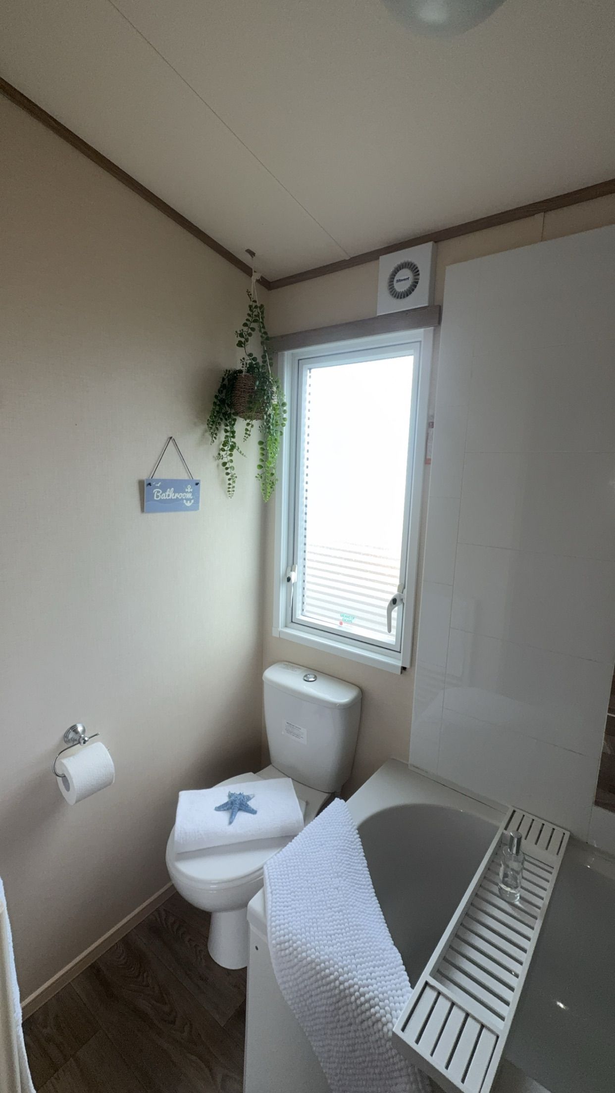 A compact bathroom with a toilet, a window, a hanging plant, and a 'Bathroom' sign.