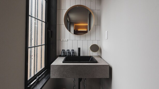 Un baño moderno con un lavamanos rectangular, un espejo redondo y una ventana con vistas a un edificio.