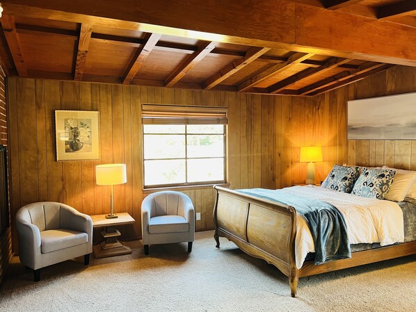 A bedroom with a wooden ceiling, a bed with patterned pillows, a chair, a bedside table, and a window with blinds.