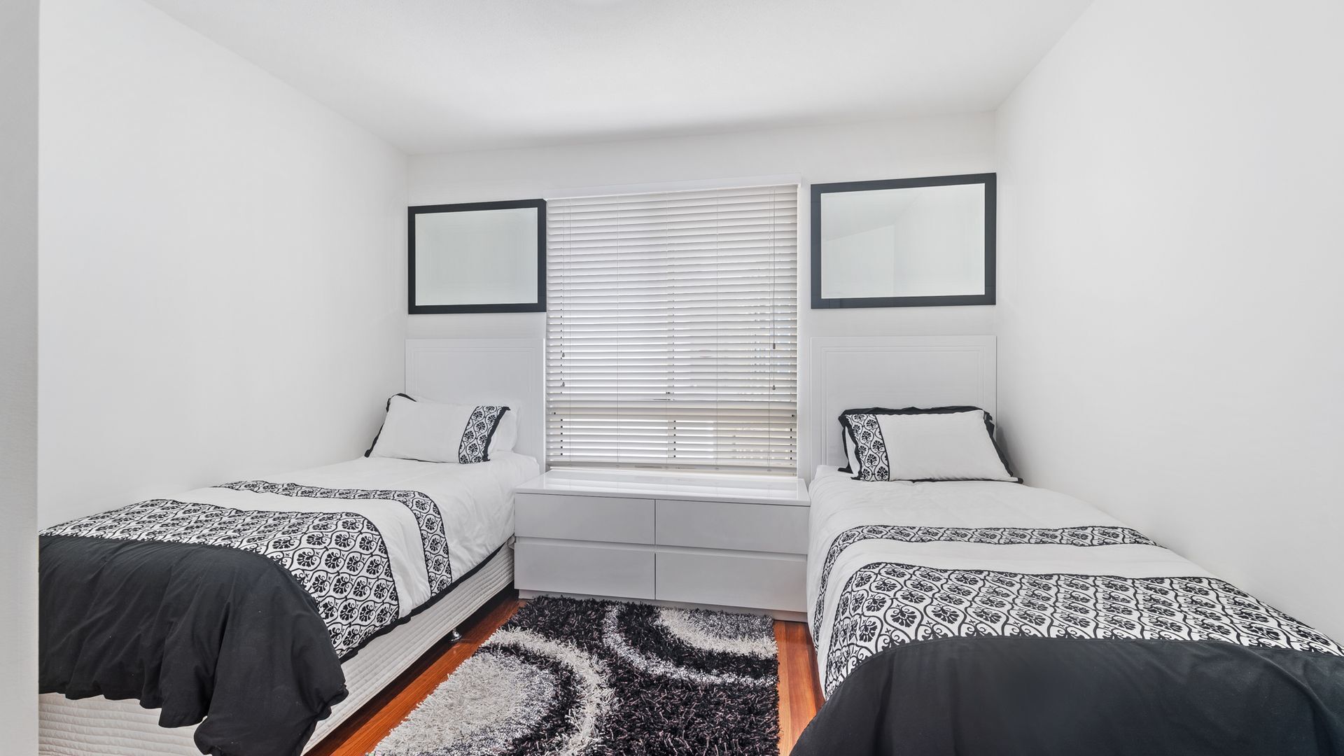 A bedroom with two beds, a window with blinds, a mirror above each bed, and a small bedside table.