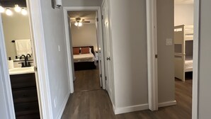 Un corridoio con pavimento in legno, pareti bianche e porte che conducono alle camere da letto. Si vede una ventola a soffitto e un lampadario.
