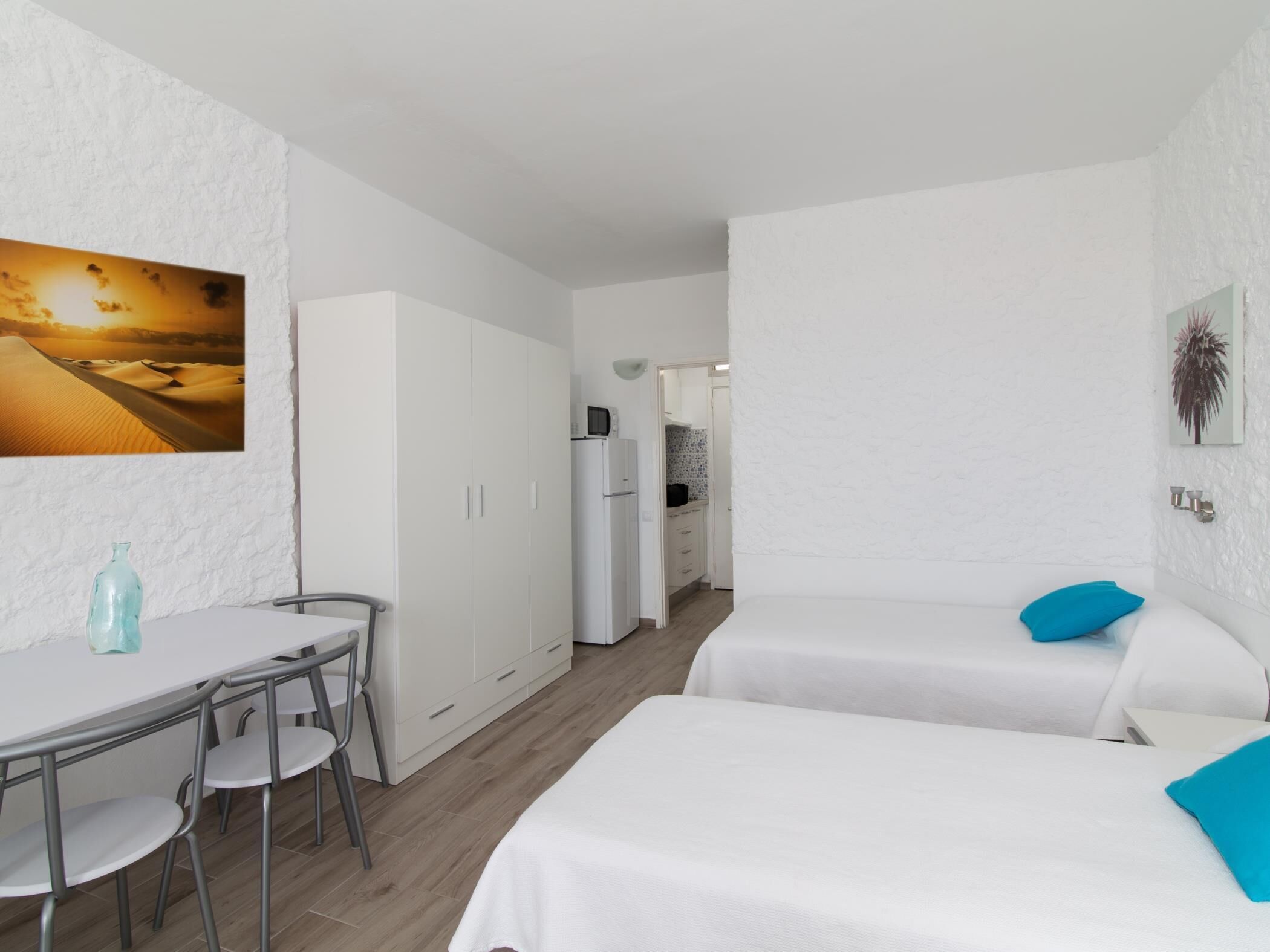 Una habitación de hotel moderna con zona de comedor, cama con almohadas azules y cocina al fondo.