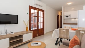 A modern living room with a flat-screen TV, wooden furniture, a dining table with chairs, and a kitchen area with white cabinets and appliances.