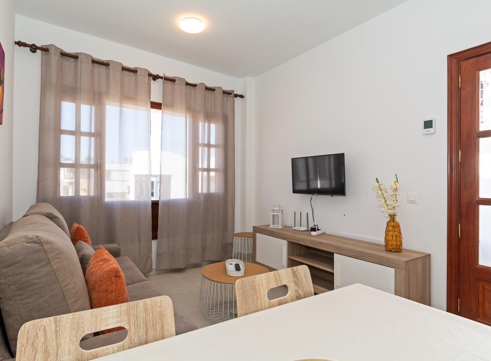 Et moderne hotelværelse med en sofa, et spisebord, et fjernsyn ophængt på væggen og et vindue med gardiner.