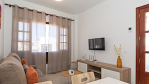 A modern hotel room with a sofa, a dining table, a TV mounted on the wall, and a window with curtains.