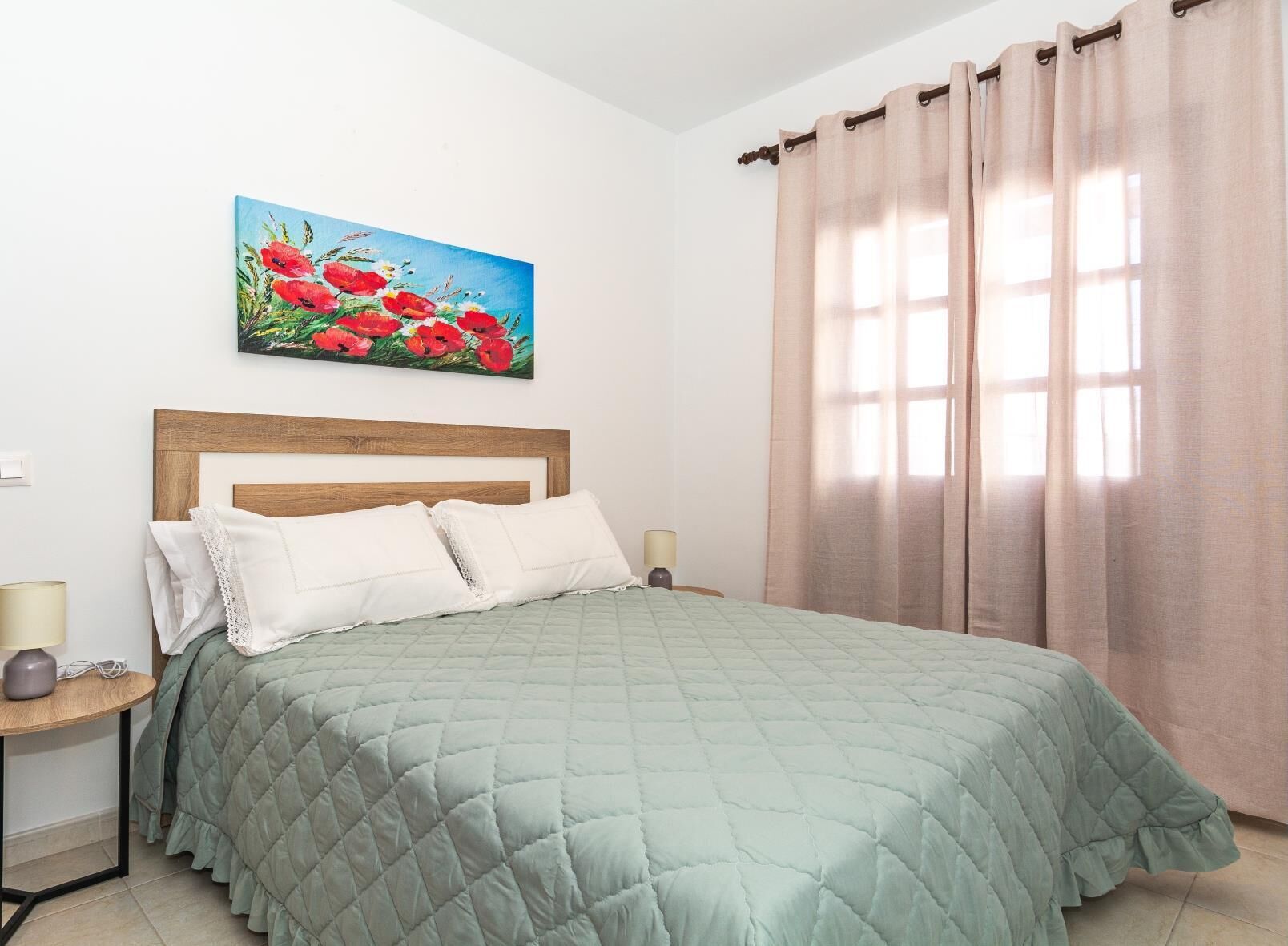Un dormitorio con cabecera de madera, colcha verde acolchada, almohadas blancas, una mesita de noche con lámpara y un cuadro de flores rojas en la pared.