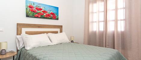 Un dormitorio con cabecera de madera, colcha verde acolchada, almohadas blancas, una mesita de noche con lámpara y un cuadro de flores rojas en la pared.