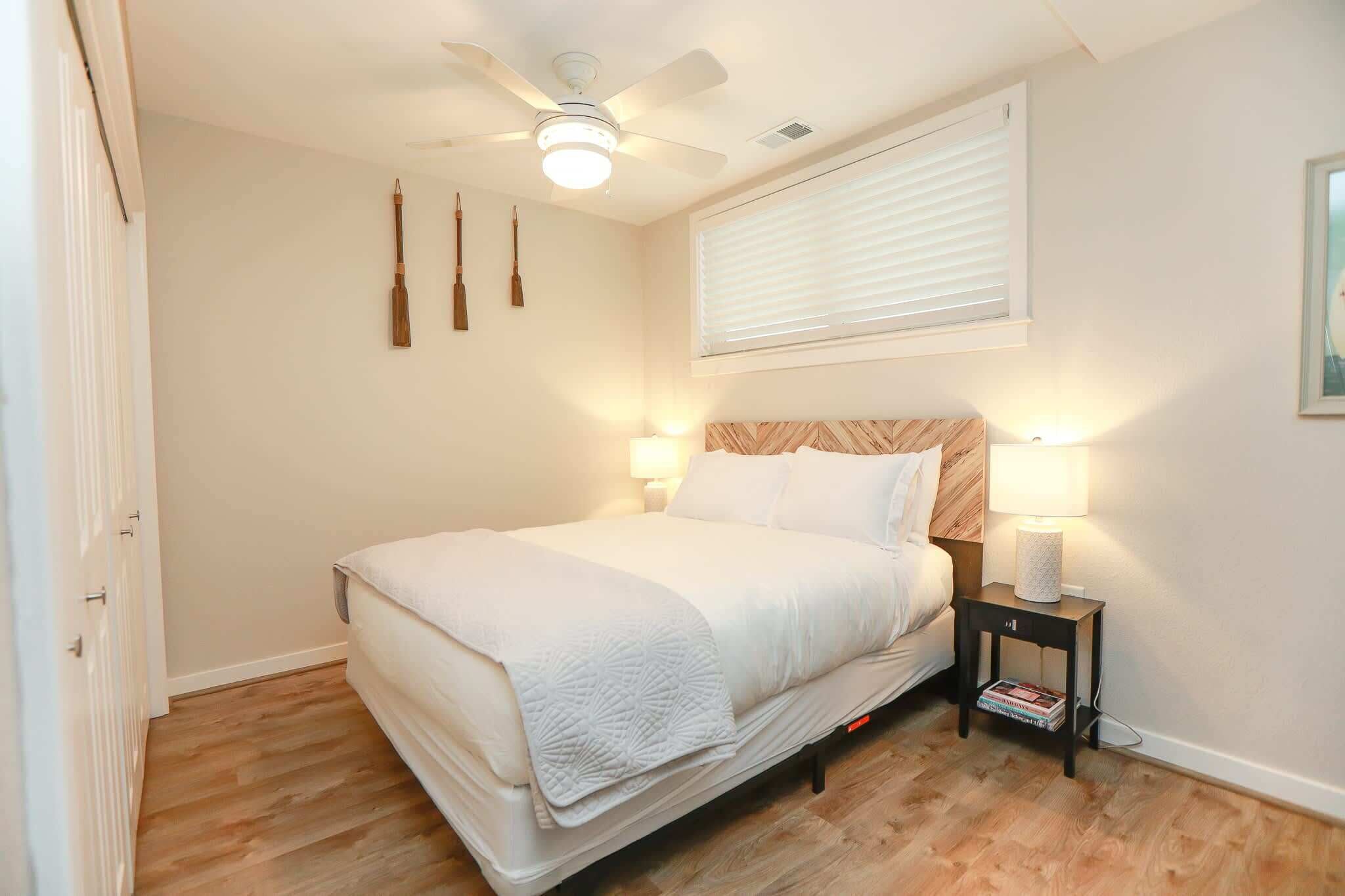 Ein Schlafzimmer mit einem Bett, einem Deckenventilator, einem Nachttisch mit Lampe und einem Fenster mit Jalousien.