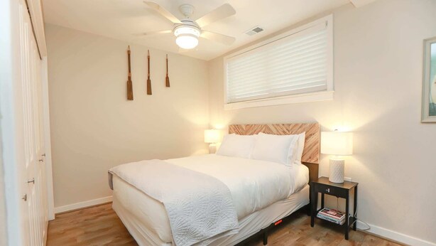 A bedroom with a bed, a ceiling fan, a nightstand with a lamp, and a window with blinds.