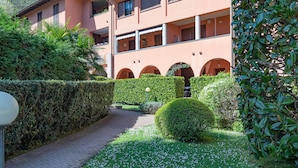 Un sendero lleva a un edificio con varios balcones, rodeado de setos cuidados y un pequeño jardín con flores blancas.