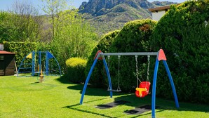 Ein Spielplatz mit Schaukeln und einer Rutsche, umgeben von grünem Gras und Bäumen, mit Bergen im Hintergrund.