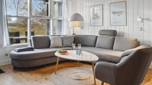 A modern living room with a grey sectional sofa, a round coffee table, and a lamp. There are two windows with curtains, a wall-mounted air conditioner, and framed artwork.
